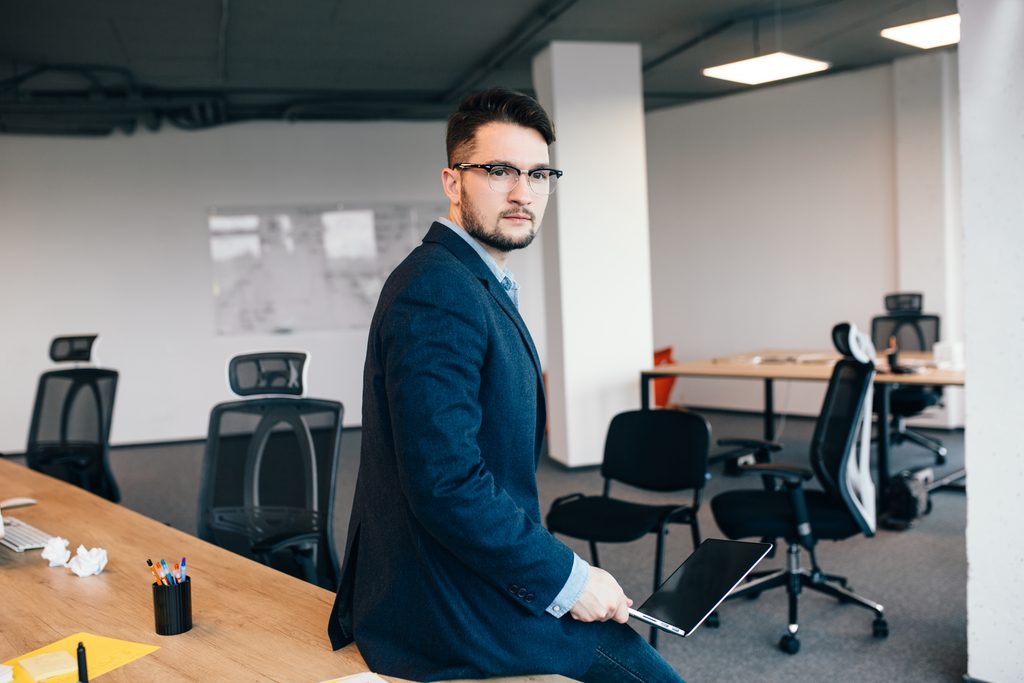 Un homme à lunettes est assis sur une table près de son poste de travail, au bureau. Il porte une chemise bleue et une veste foncée. Il a l'air sérieux.